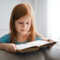 little girl with red hair reading a book on a sofa